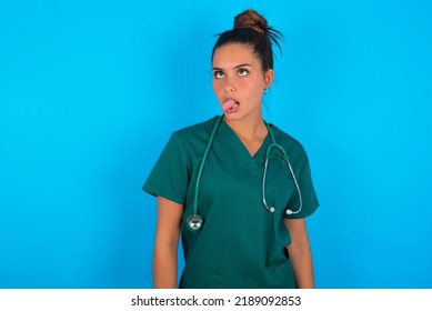 Beautiful Doctor Woman Wearing Medical Uniform Over Blue Background Showing Grimace Face Crossing Eyes And Showing Tongue. Being Funny And Crazy