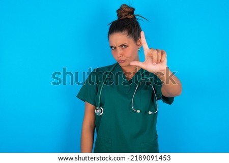 beautiful doctor woman wearing medical uniform over blue background making fun of people with fingers on forehead doing loser gesture mocking and insulting.