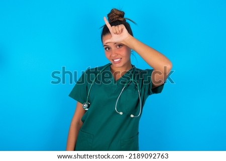 beautiful doctor woman wearing medical uniform over blue background making fun of people with fingers on forehead doing loser gesture mocking and insulting.