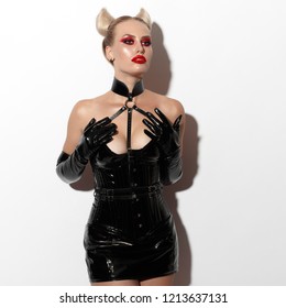 Beautiful dominant blonde devil demon vamp mistress bdsm girl with hair horns in glamour latex skirt, corset, collar and bdsm black leather fetish harness posing on white background