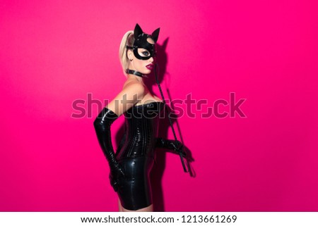 Beautiful dominant blonde vamp mistress bdsm girl with fashion makeup in glamour latex skirt, corset, collar and bdsm black leather fetish cat mask posing with whip on hot pink background