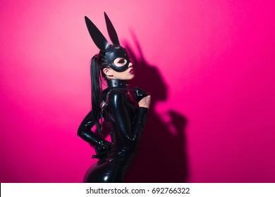 Beautiful dominant brunette vamp mistress girl with fashion makeup in glamour latex dress, collar and bdsm black leather fetish rabbit mask posing on hot pink backgroung