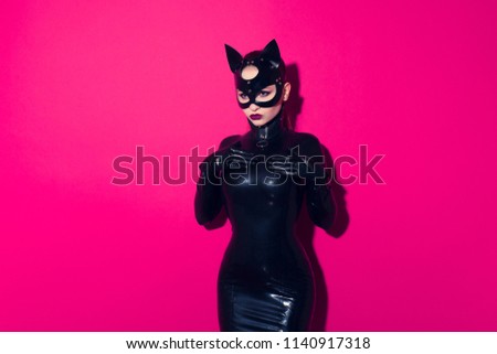 Beautiful dominant brunette vamp mistress bdsm girl with fashion makeup in glamour latex dress, collar and black leather fetish cat mask posing on hot pink background