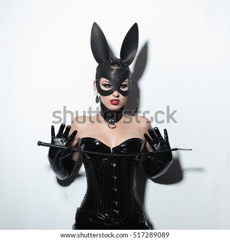 Beautiful dominant brunette vamp mistress girl in latex corset, long gloves, collar and bdsm black leather fetish rabbit mask posing with riding crop on white backgroung