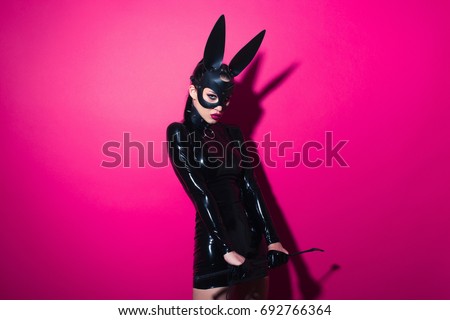 Beautiful dominant brunette vamp mistress girl with fashion makeup in glamour latex dress, collar and bdsm black leather fetish rabbit mask posing on hot pink backgroung