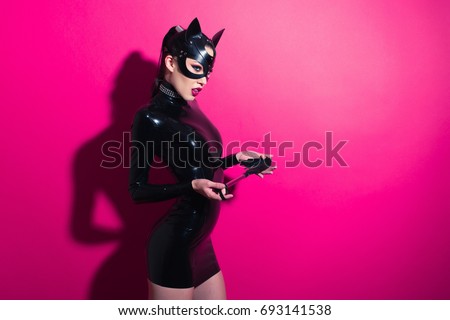 Beautiful dominant brunette vamp mistress girl with fashion makeup in glamour latex dress, collar and bdsm black leather fetish cat mask posing on hot pink backgroung