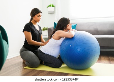 Beautiful doula in activewear helping a caucasian pregnant woman with her back pain. Expectant mother relaxing in a fitness ball while receiving a massage