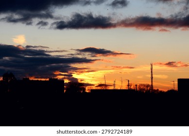 Beautiful dramatic sunset with dark clouds over the city silhouette. Bright orange and yellow sun light breaking through the evening sky. Urban landscape at dusk with industrial details and tall antenna.