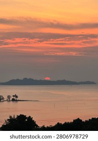 Beautiful dramatic sunset over the ocean with a red sun setting behind a distant island silhouette and orange cloudy sky.