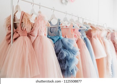 Beautiful dressy lush pink and blue dresses for girls on hangers at the background of white wall. Kids dresses with feathers for prom and holiday.
