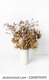 Beautiful dried flower arrangement of Australian native banksia, eucalyptus leaves, red leucadendrons and delicate white flowers, in a white vase on a table with a white background.