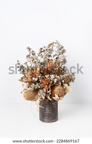 Beautiful dried flower arrangement of Australian native banksia, eucalyptus leaves, red leucadendrons and delicate white flowers, in a glass vase on a table with a white background.