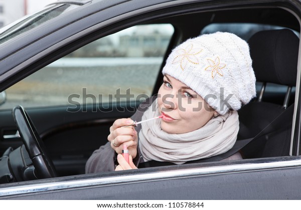 Beautiful driver in winter clothes in car applying\
make up