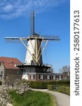 A beautiful Dutch windmill with large blades towers over a charming village, surrounded by blooming trees and a clear blue sky, creating an idyllic and quintessentially Dutch scene.