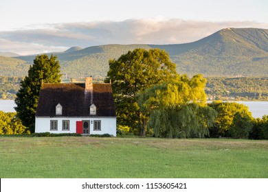 Beautiful Early Morning Summer View Of Patrimonial White Wooden House With Steep Shingled Roof In Rural Setting With Trees, River And Mountains In The Background, Island Of Orleans, Quebec, Canada