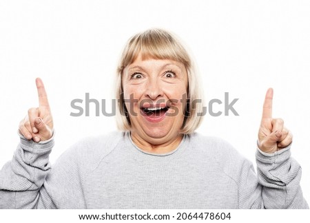 Beautiful elderly female model with optimistic expression indicates upwards, being excited by something, raises fore finger, poses against white background with copy space for your promotional text