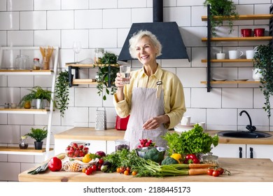 beautiful elderly gray haired senior woman cook in cozy kitchen with fresh organic vegetables on table for healthy vegetable salad, drink pure clear water from glass , healthy food for active life - Powered by Shutterstock