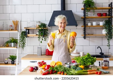 Beautiful Elderly Gray Haired Senior Woman Cook In Cozy Kitchen With Fresh Organic Vegetables, Making  Fresh Orange Juice On Table And Cooking Healthy Vegetable Salad, Healthy Food Active Life