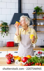 Beautiful Elderly Gray Haired Senior Woman Cook In Cozy Kitchen With Fresh Organic Vegetables, Making  Fresh Orange Juice On Table And Cooking Healthy Vegetable Salad, Healthy Food Active Life