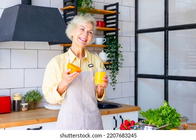 Beautiful Elderly Gray Haired Senior Woman Cook In Cozy Kitchen With Fresh Organic Vegetables, Making  Fresh Orange Juice On Table And Cooking Healthy Vegetable Salad, Healthy Food Active Life