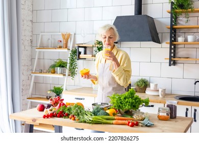 Beautiful Elderly Gray Haired Senior Woman Cook In Cozy Kitchen With Fresh Organic Vegetables, Making  Fresh Orange Juice On Table And Cooking Healthy Vegetable Salad, Healthy Food Active Life