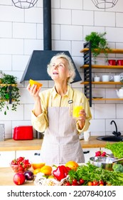 Beautiful Elderly Gray Haired Senior Woman Cook In Cozy Kitchen With Fresh Organic Vegetables, Making  Fresh Orange Juice On Table And Cooking Healthy Vegetable Salad, Healthy Food Active Life