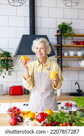 Beautiful Elderly Gray Haired Senior Woman Cook In Cozy Kitchen With Fresh Organic Vegetables, Making  Fresh Orange Juice On Table And Cooking Healthy Vegetable Salad, Healthy Food Active Life