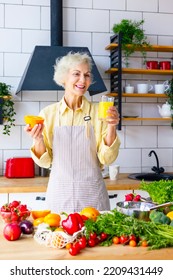 Beautiful Elderly Gray Haired Senior Woman Cook In Cozy Kitchen With Fresh Organic Vegetables, Making  Fresh Orange Juice On Table And Cooking Healthy Vegetable Salad, Healthy Food Active Life