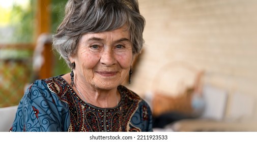 A beautiful elderly woman aged 80+ with a smiling face sits on the veranda of her cozy house in a smart dress, a happy pensioner takes care of her beauty and health.