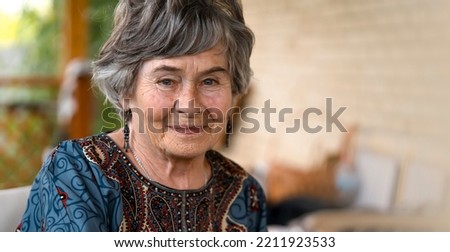 A beautiful elderly woman aged 80+ with a smiling face sits on the veranda of her cozy house in a smart dress, a happy pensioner takes care of her beauty and health.