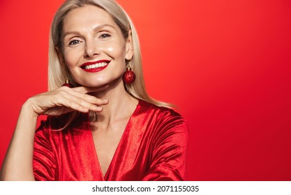 Beautiful elderly woman in evening dress, red lipsticks and Christmas tree bubble earrings, smiling at camera, touching her soft, clear facial skin, red background - Powered by Shutterstock