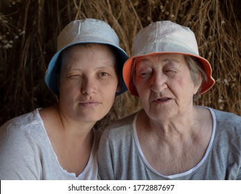 Beautiful Elderly Woman With Wrinkles And Young Girl From Same Family. Two Different Ages Of Similar People. Mother And Adult Daughter In Identical Hats On Background Of Dried Flowers