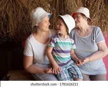 Beautiful Elderly Woman With Wrinkles, Young Girl And Boy From Same Family. Three Different Ages Of Similar People. Mother, Adult Daughter And Grandson In Identical Hats On Background Of Dry Flowers