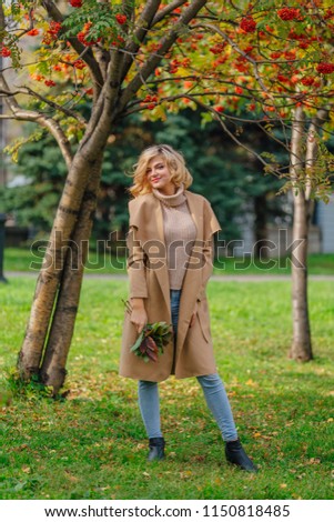 Beautiful elegant blonde woman dressed in a coat standing under the rowan tree holding bouquet of rowanberries and leaves in autumn park. Sunny october day.