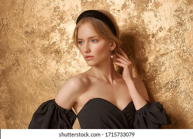 Beautiful Elegant Blonde Woman With Hairstyle Wearing Casual Clothes, Black Top And White Trousers With Band In Hair Standing Leaning On Golden Wall And Posing. Styish Fashion Female Model