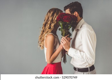 Beautiful Elegant Couple Is Kissing Behind Roses, On Gray Background