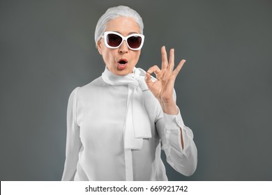Beautiful elegant elderly woman with sunglasses showing ok gesture on gray background