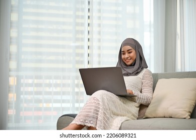 Beautiful and elegant millennial Asian Muslim woman or businesswoman with hijab using laptop computer to manage her tasks while relaxing on her comfortable sofa in the living room. work from home - Powered by Shutterstock