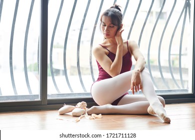 Beautiful elegant pensive dancer in leotard and ballet slippers resting on floor next to big window after training and touching face
