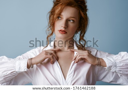 Beautiful elegant redhead freckled woman wearing luxury silver jewelry: earrings, chain, bracelets, white shirt, posing in studio, on blue background. Jewellery advertising concept. Close up portrait