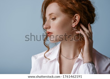 Beautiful elegant redhead freckled woman wearing luxury silver jewelry: earrings, chain, posing in studio, on blue background. Jewellery advertising conception. Close up profile portrait. Copy space