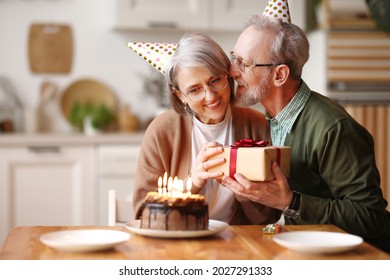 Beautiful elegant senior couple celebrating birthday together at home, loving elderly man congratulating woman wife, giving present gift box and kissing gently in cheek, cake with candles on table - Shutterstock ID 2027291333