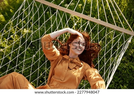 a beautiful, elegant woman lies in a long orange dress on a mesh hammock resting in nature, illuminated by the warm sunset light, holding her hand on her head, covering her face from the sun