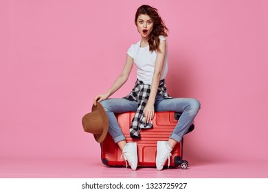 Beautiful elegant woman sitting on a red suitcase with a hat in hand pink travel background