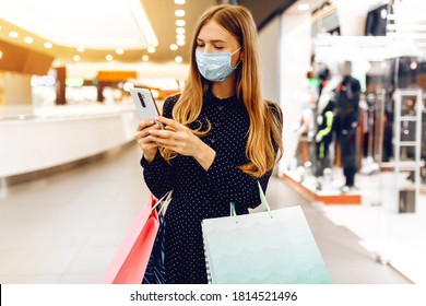 beautiful elegant young woman in a medical protective mask on her face, with shopping bags, uses a mobile phone while in a shopping center