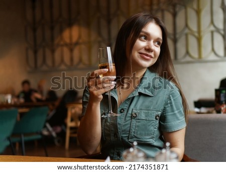 Beautiful embarassed female with cheerful expression sitting with glass of wine in the cafe while waiting for the meal. Lifestyle, relax, chill, dinner, supper, meal