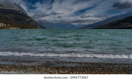 The beautiful emerald lake is surrounded by mountains. Ripples on the water, foam of waves on the pebbly shore. Snow-capped mountain peaks of the Andes against a  blue sky and clouds. Lago Roca. - Powered by Shutterstock