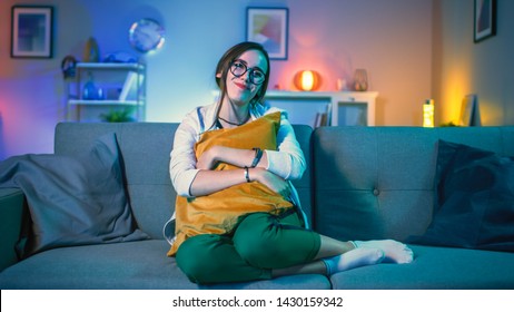 Beautiful Emotional Young Girl In Glasses Sitting On A Couch And Watching TV At Home. She Is Hugging A Pillow. Screen Adds Reflections To Her Face. Cozy Room Is Lit With Warm Light.