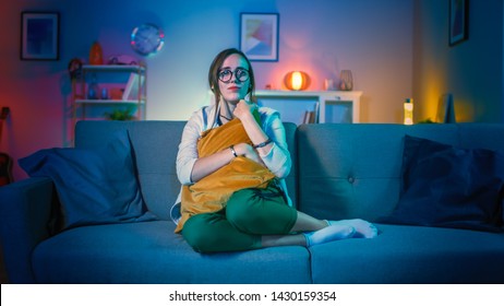 Beautiful Emotional Young Girl In Glasses Sitting On A Couch And Watching TV At Home. She Is Hugging A Pillow. Screen Adds Reflections To Her Face. Cozy Room Is Lit With Soft Light.