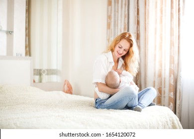 Beautiful Emotional Young Mom Blonde With Long Hair Breastfeeds Her Baby In A Bright Bedroom, Pretty Mother In Jeans Breastfeeds Her Baby 6 Months Old, Concept Of Motherhood And Breastfeeding